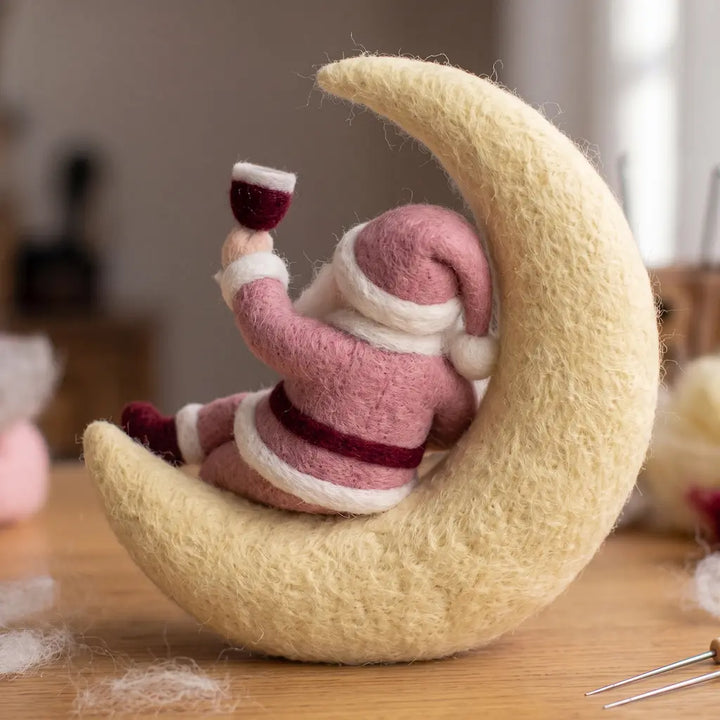Back view of a pink felt Santa seated on a cream crescent moon, showing his drooping hat, burgundy belt and raised goblet.