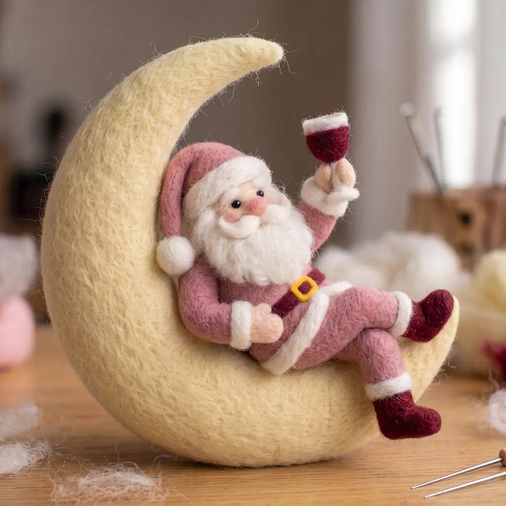 Pink wool felt Santa reclining on a cream crescent moon, raising a burgundy goblet with his legs crossed.