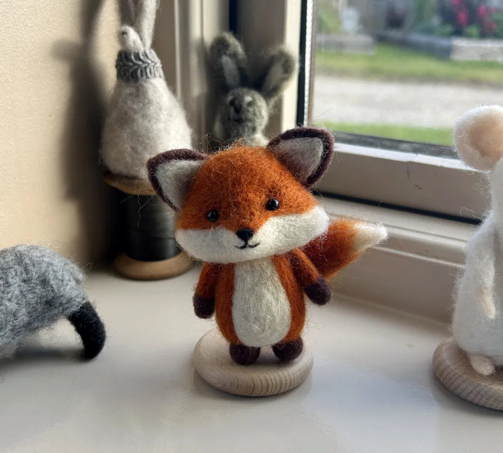 An orange felt fox on a wooden base, displayed among other felt animals on a windowsill.