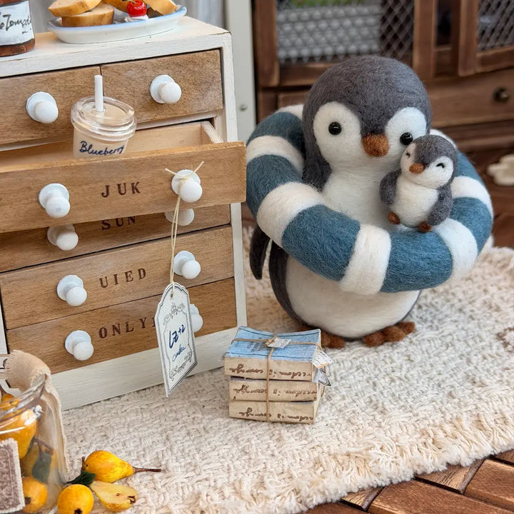 A felt penguin figurine holding a smaller penguin, styled on a cream rug beside a wooden drawer cabinet with decorative knobs and tags.