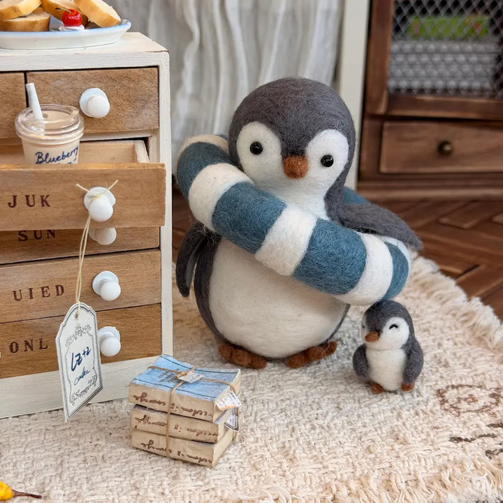 A felt penguin plush figurine with its smaller matching penguin figure, styled on a textured rug next to a wooden drawer chest and a small wrapped gift box.