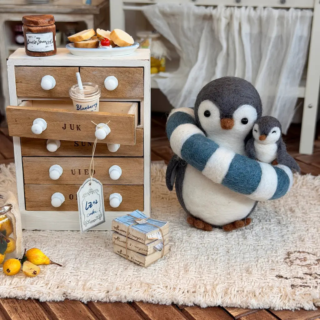 A felt penguin mother holding a small penguin chick, styled in a cozy miniature room scene next to a wooden drawer side table with tiny props.