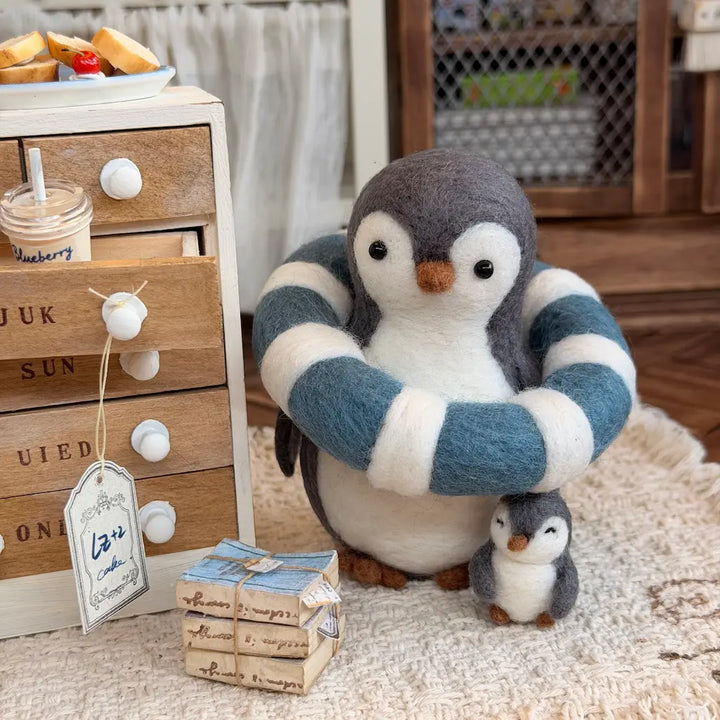 A felt penguin figurine with a blue-and-white striped scarf stands on a cream rug beside a small matching penguin figure. The scene includes a wooden drawer cabinet, a drink, and a small gift box in a styled indoor setting.
