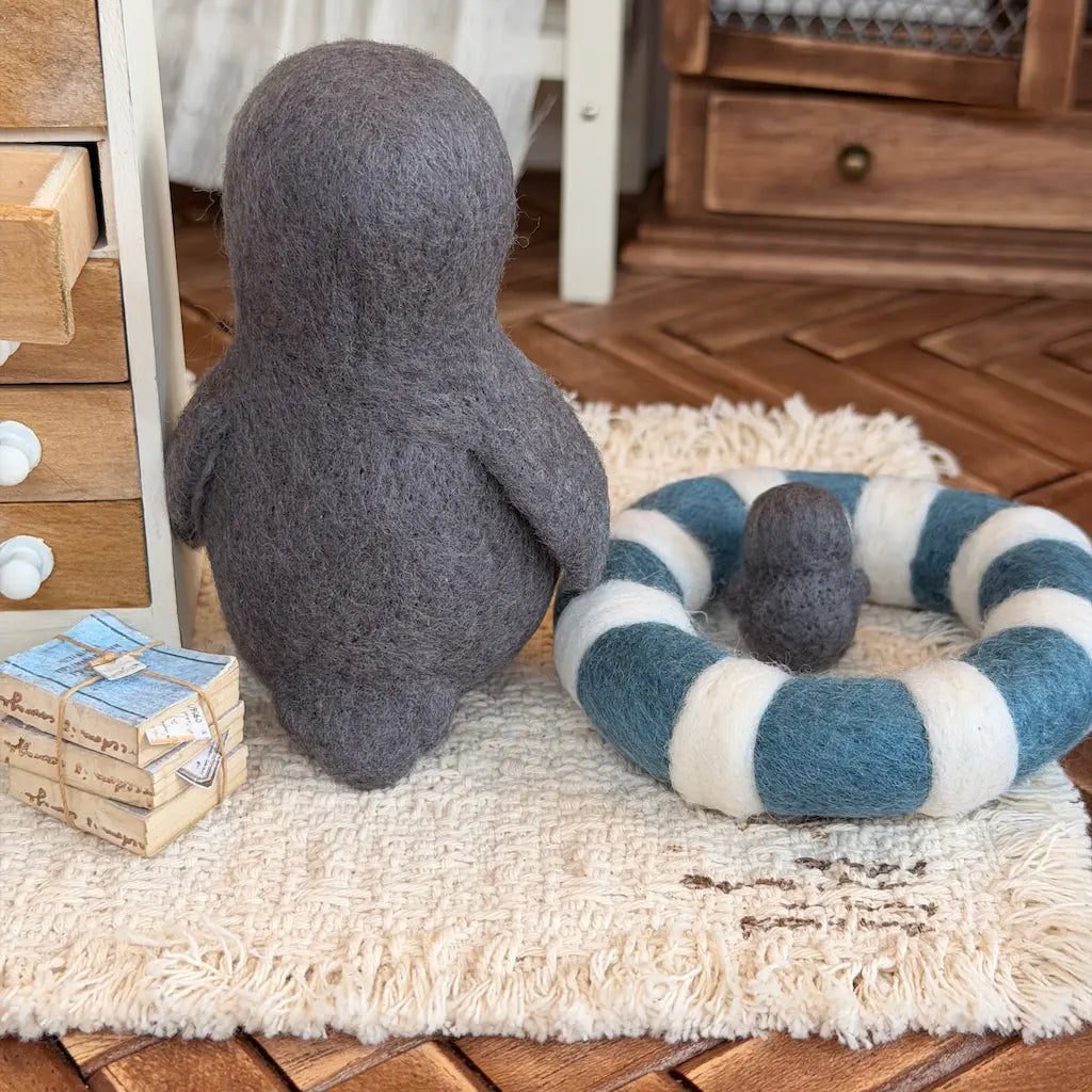 A felt penguin figurine in dark gray sits on a cream rug next to a blue and white striped felt ring cushion with a small matching gray ball, in a styled indoor setting with wooden furniture.