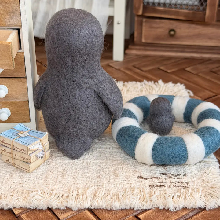 A felt penguin figurine in dark gray sits on a cream rug next to a blue and white striped felt ring cushion with a small matching gray ball, in a styled indoor setting with wooden furniture.