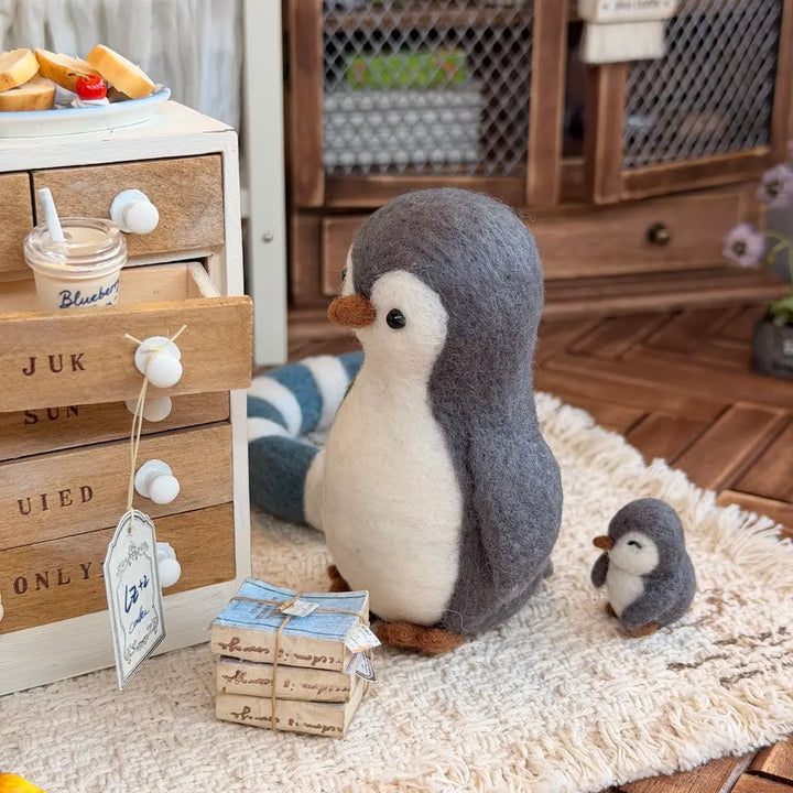 A felt penguin plush figurine sits on a cream rug beside a small matching penguin figure, with a wooden drawer unit and cozy room decor in the background.