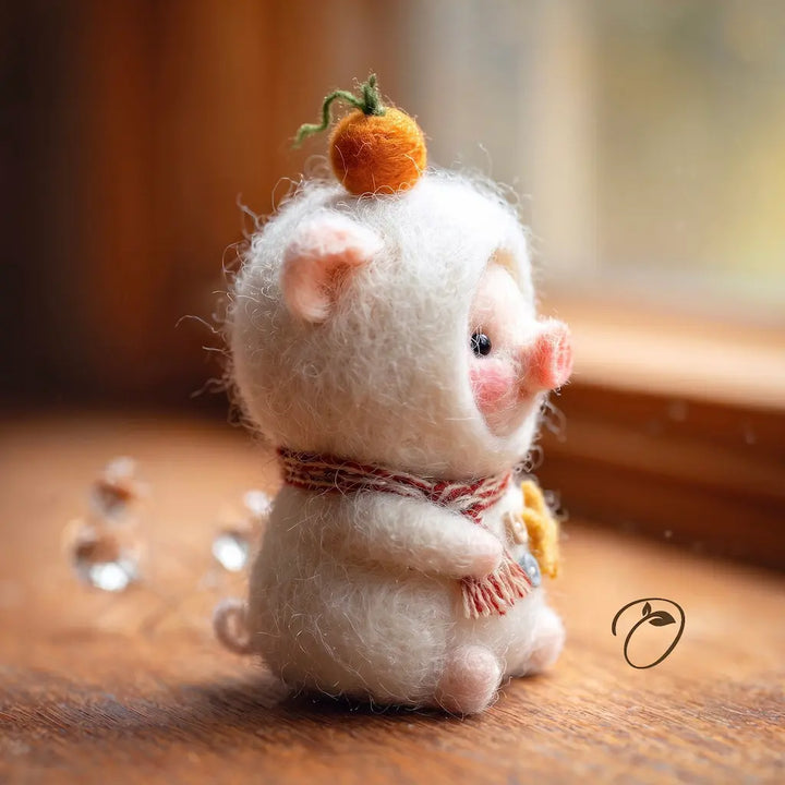 A small felted wool pig figurine with pink ears and snout, wearing a red-and-white striped scarf and holding a yellow flower, sitting on a wooden surface near a window.
