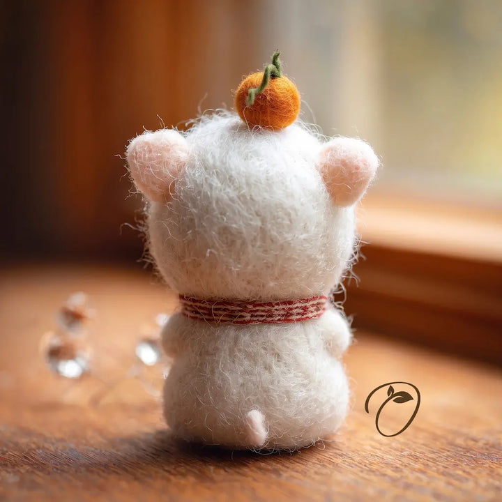 A small white felted-fur animal figurine shown from the back on a wooden surface, with pink ears, a red-and-white striped band around its neck, and an orange pom-pom on top. The background is softly blurred with warm light coming from a window.