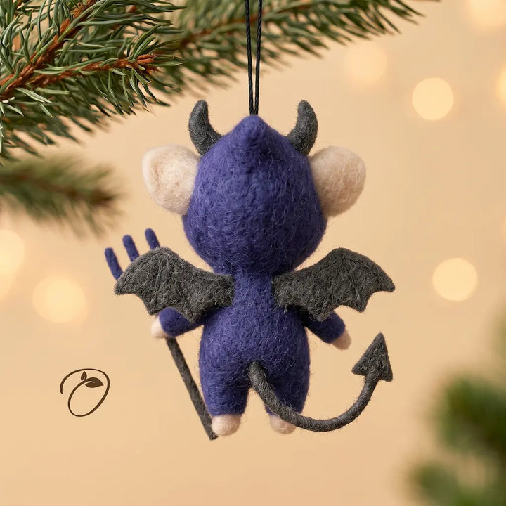 A felt Christmas ornament shaped like a small blue-purple devil hanging from a black string, with gray wings, tail, and staff, photographed against a warm blurred holiday background with pine branches at the top.