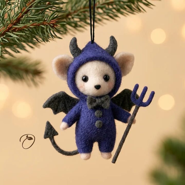 A felt Christmas ornament shaped like a small devil mouse wearing a purple hooded outfit, with black wings, a black tail, and holding a black pitchfork, hanging against a warm blurred holiday background with pine branches at the top.