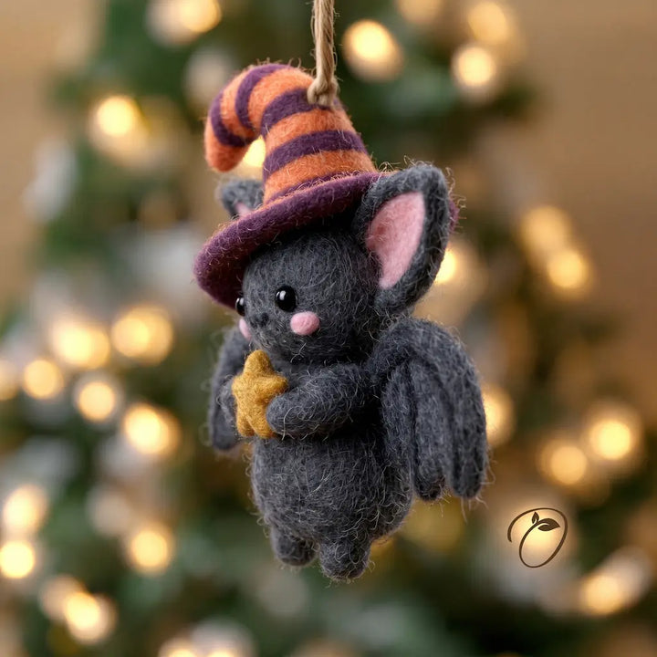 A small felted wool bat hanging ornament wearing an orange and purple striped witch hat and holding a yellow star, set against a blurred Christmas tree with warm lights.