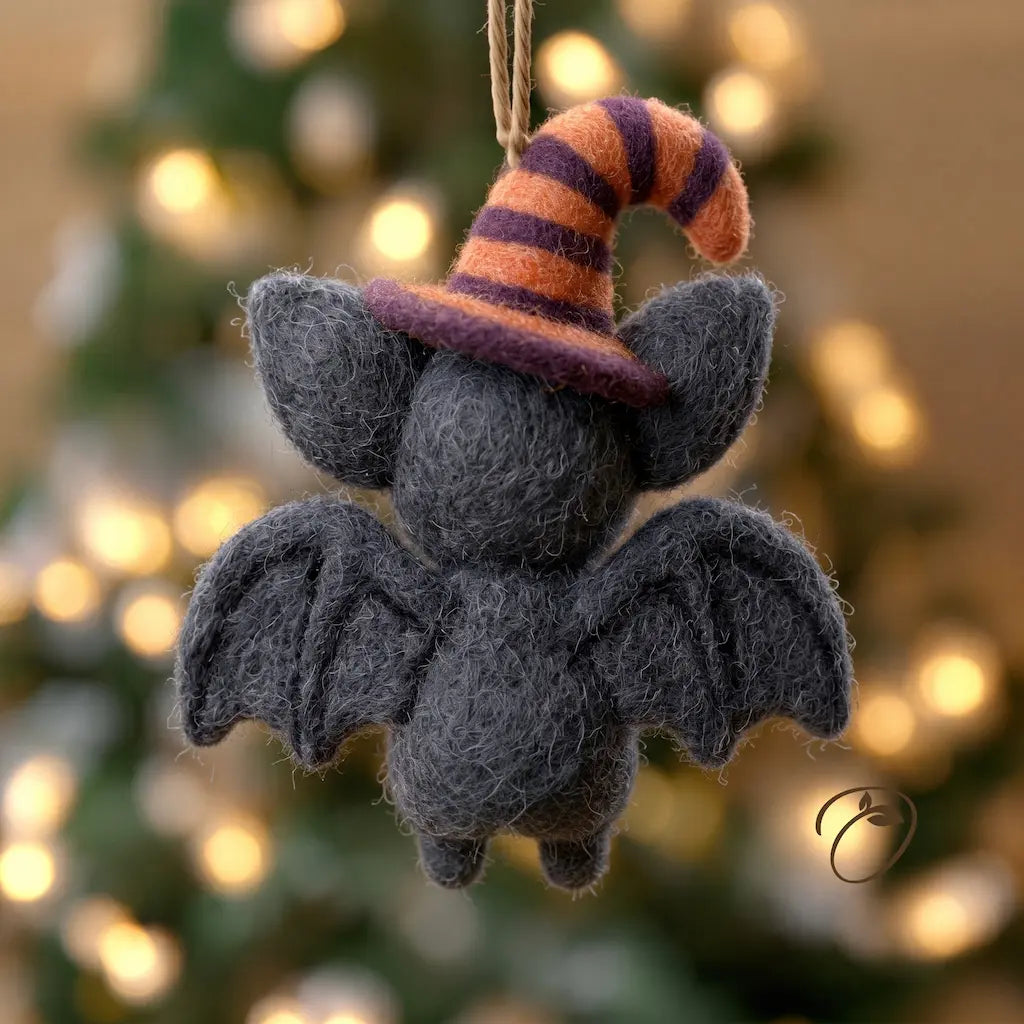 A dark gray felted wool bat hanging ornament with a purple and orange striped witch hat, suspended by a string against a blurred Christmas tree background with warm lights.