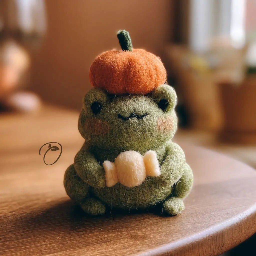A cute handmade felt plush frog holding a cream-colored candy, wearing an orange felt pumpkin hat with a green stem. The plush frog is sitting on a warm wooden table with a soft, blurred background.