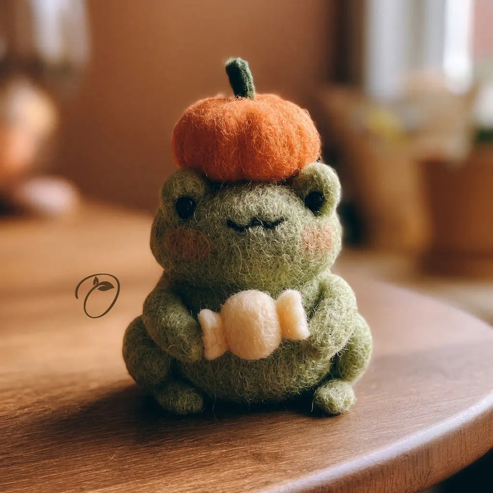 A cute handmade felt plush frog holding a cream-colored candy, wearing an orange felt pumpkin hat with a green stem. The plush frog is sitting on a warm wooden table with a soft, blurred background.