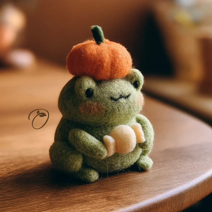 A cute felted wool frog decoration sitting on a wooden table, wearing an orange felted pumpkin hat and holding a cream-colored acorn.