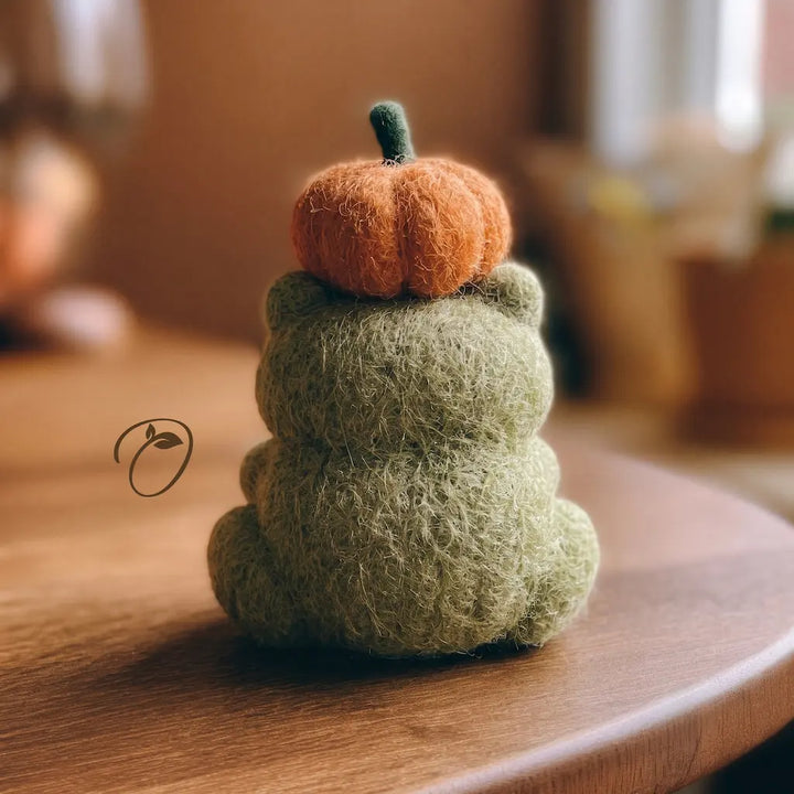 A small felted wool decorative figure shaped like a seated figure, with an orange pumpkin on its head, photographed on a wooden table in a softly blurred indoor setting.