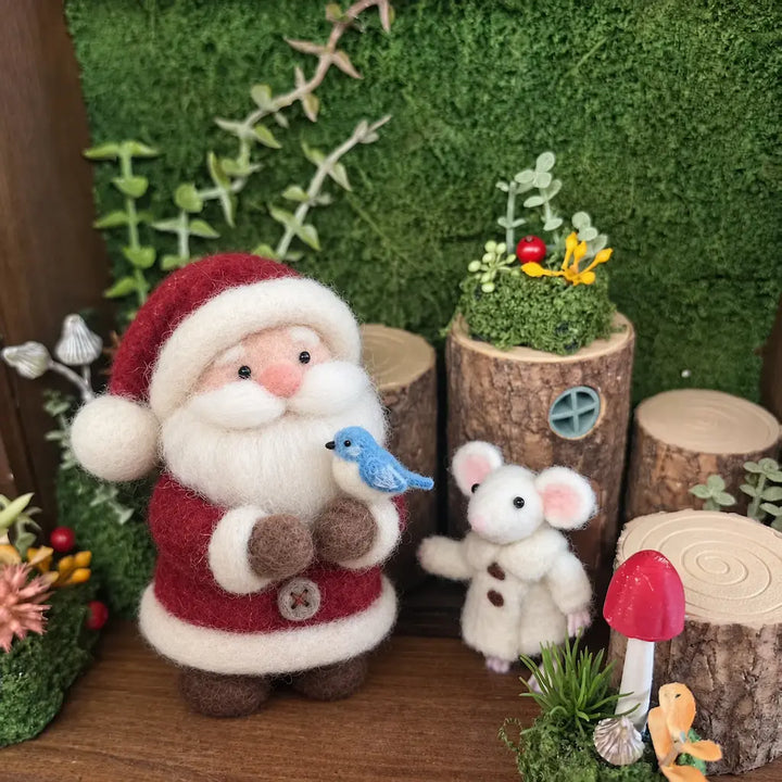A festive miniature felt decoration set featuring a Santa Claus figure in red and white holding a blue bird, next to a small white mouse in a cozy woodland scene with faux moss, wood logs, a red mushroom, and greenery.