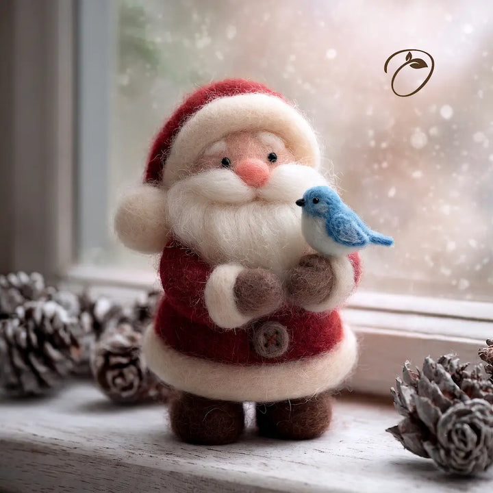 A felt Christmas ornament shaped like Santa Claus standing on a windowsill, holding a small blue bird. The ornament has a red and white outfit with brown details, surrounded by pinecones, with a soft-focus snowy window background.