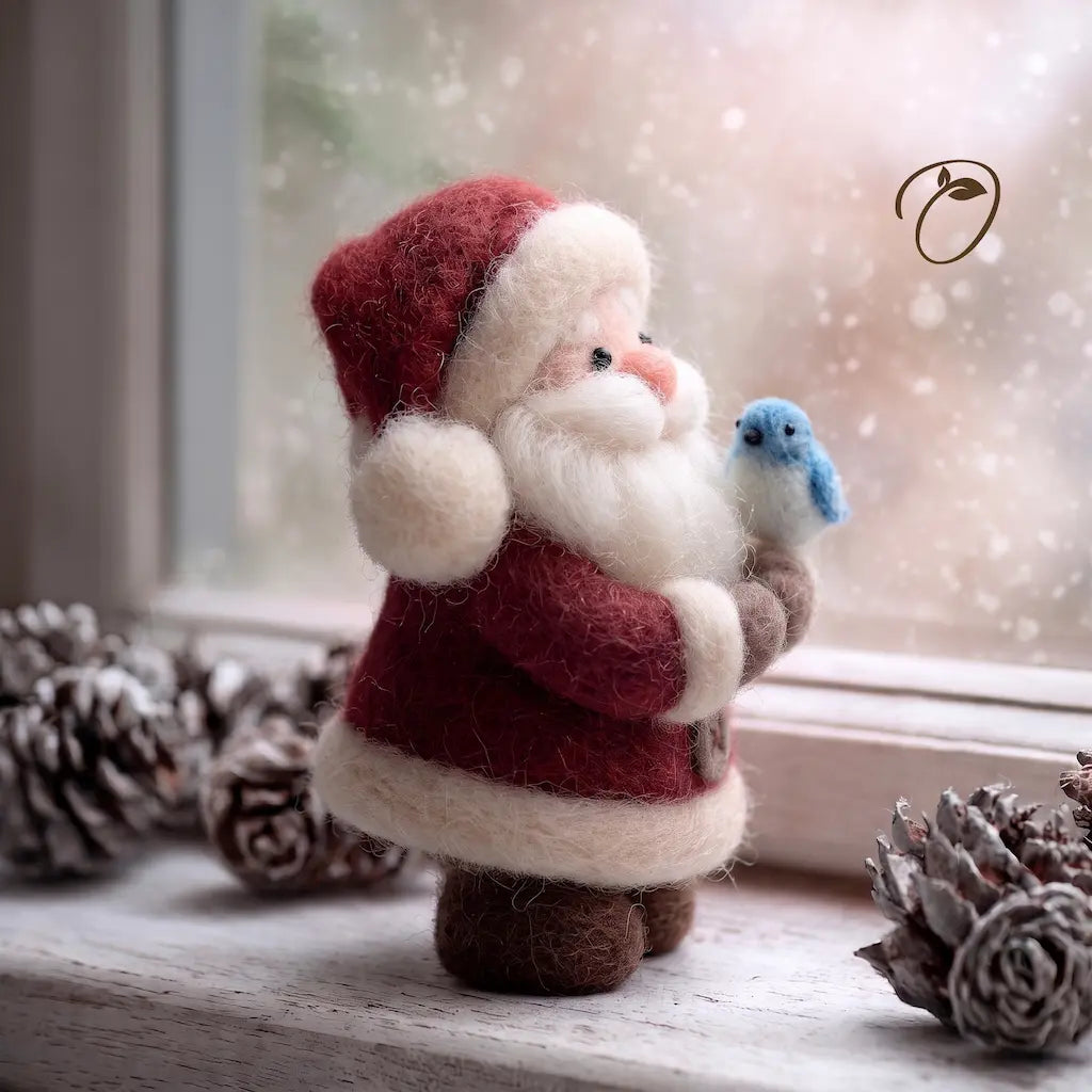 A felt Christmas ornament shaped like Santa Claus in a red suit with white trim, holding a small blue bird, displayed on a windowsill with pinecones and a snowy outdoor background.