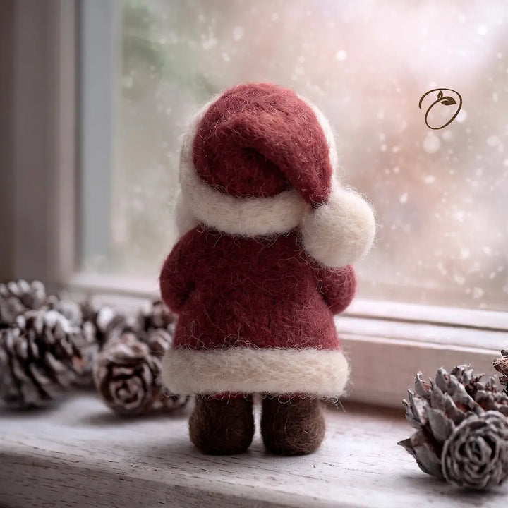 A small felted wool Santa Claus figurine seen from behind, wearing a red suit and white trim, standing on a windowsill with pinecones around it and a softly blurred snowy outdoor scene outside.