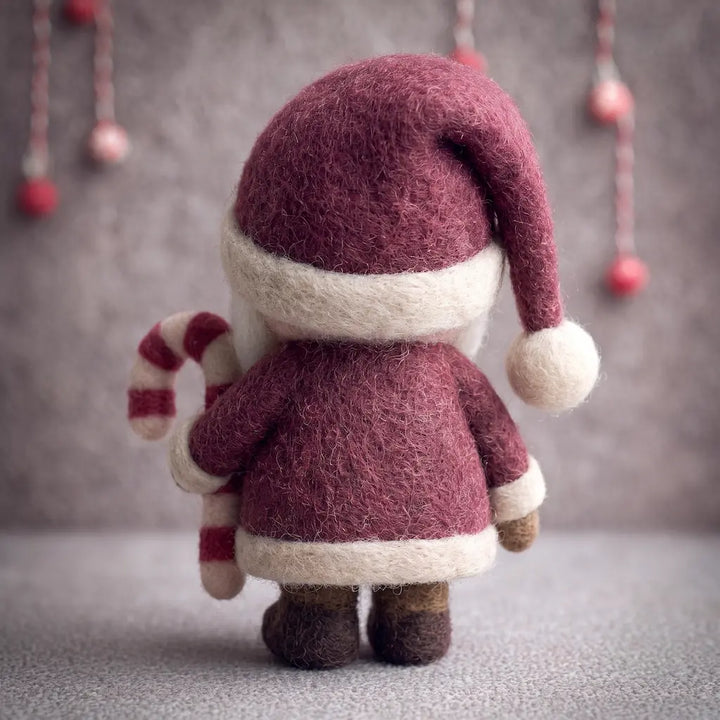 A felted wool Santa Claus figurine shown from the back, holding a striped candy cane. The Santa has a burgundy wool suit and hat with cream trim, and stands on a gray surface against a gray background with hanging decorative lights.