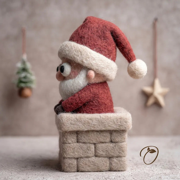 Handmade felted wool Santa Claus figurine sitting in a chimney stack, featuring a red hat with white trim, a white beard, and a red suit, set against a warm neutral background with hanging ornaments.