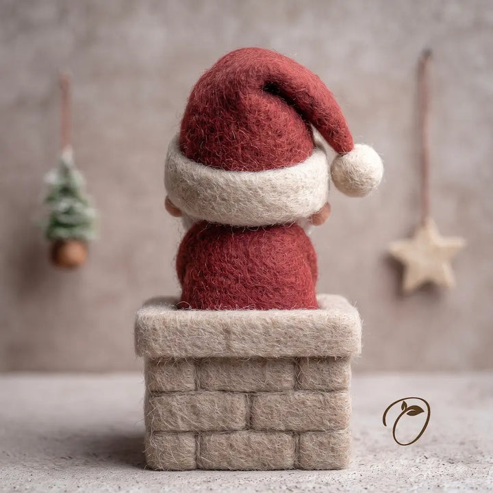 A small felted wool Christmas gnome figurine, viewed from behind, wearing a red Santa hat and red suit while sitting on a beige brick chimney. The background is a soft, textured neutral gray with hanging holiday decorations.