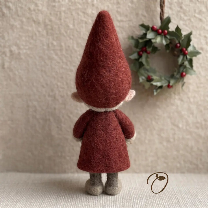 A small felt gnome figurine shown from the back, wearing a rust-red pointed hat and matching coat with light beige trim and gray shoes. It stands on a textured cream surface in front of a neutral wall, with a green holly wreath hanging in the background.