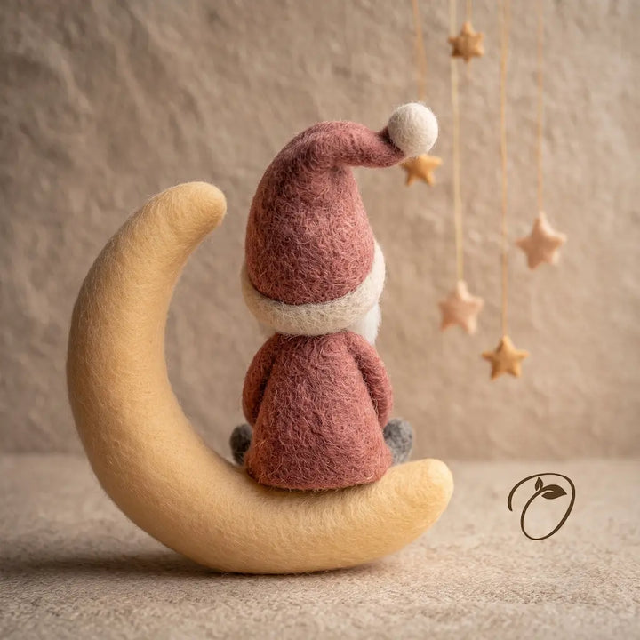 A felt Christmas ornament depicting a Santa Claus figure sitting on a crescent moon, holding a small gift box. The ornament is styled on a textured beige surface with hanging wooden star decorations in the background.