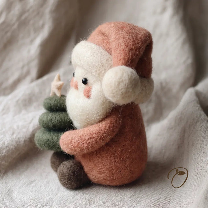A handcrafted needlepoint felt Santa Claus figurine holding a small green Christmas tree, resting on a soft beige fabric background.