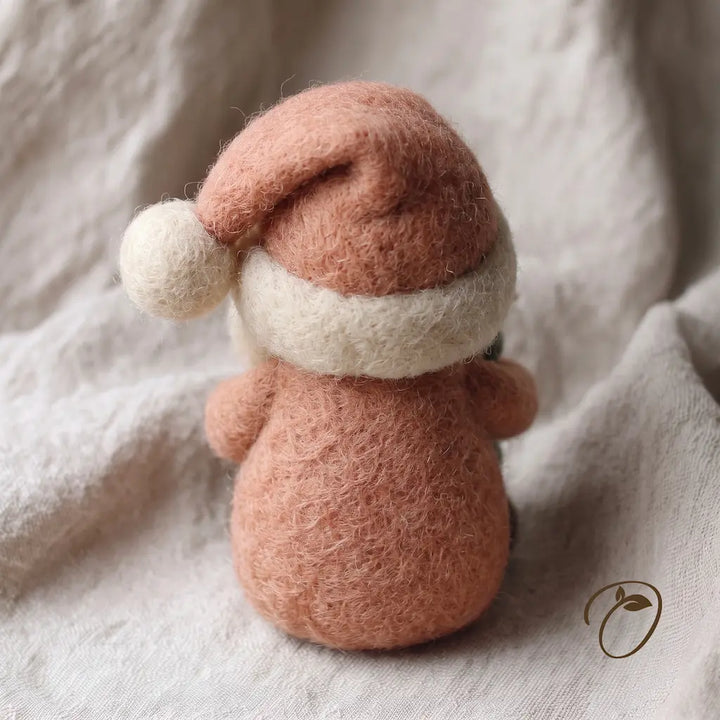 A small felted wool Christmas ornament shaped like a gingerbread man, wearing a dusty pink hat with a cream pom-pom and a cream scarf, photographed on soft beige fabric.