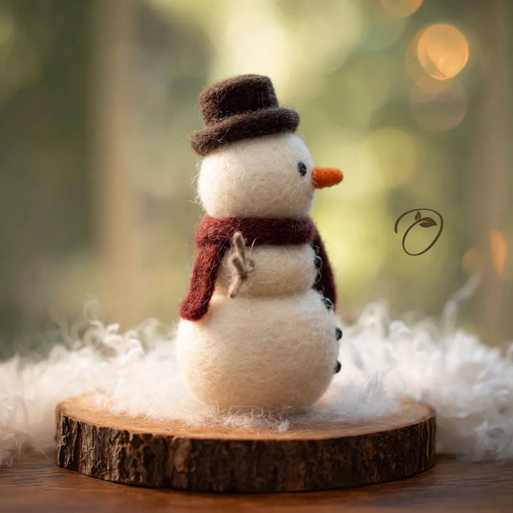 A small felt snowman ornament wearing a dark brown top hat and a burgundy scarf, standing on a round wood slice with white fluffy material around it. The background is softly blurred with warm bokeh lights.
