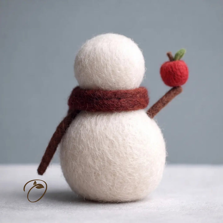 A small felt snowman decoration with a white body and hat, a dark red scarf, brown arms, and a red apple accessory, photographed against a soft gray background.