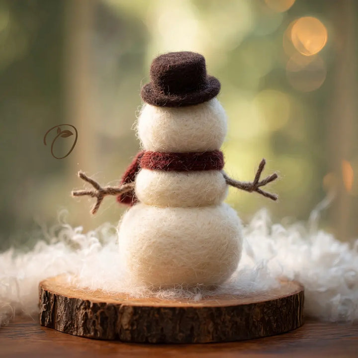 A small felt snowman decoration with a dark brown top hat, cream-colored body, a dark red scarf, and twig arms, displayed on a round wood slice with fluffy white fiber around it against a softly blurred outdoor background.