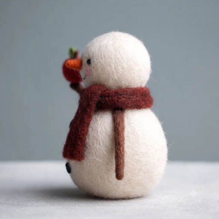A small felt snowman decoration with a white body, dark red scarf, and orange carrot nose, standing against a soft gray background.