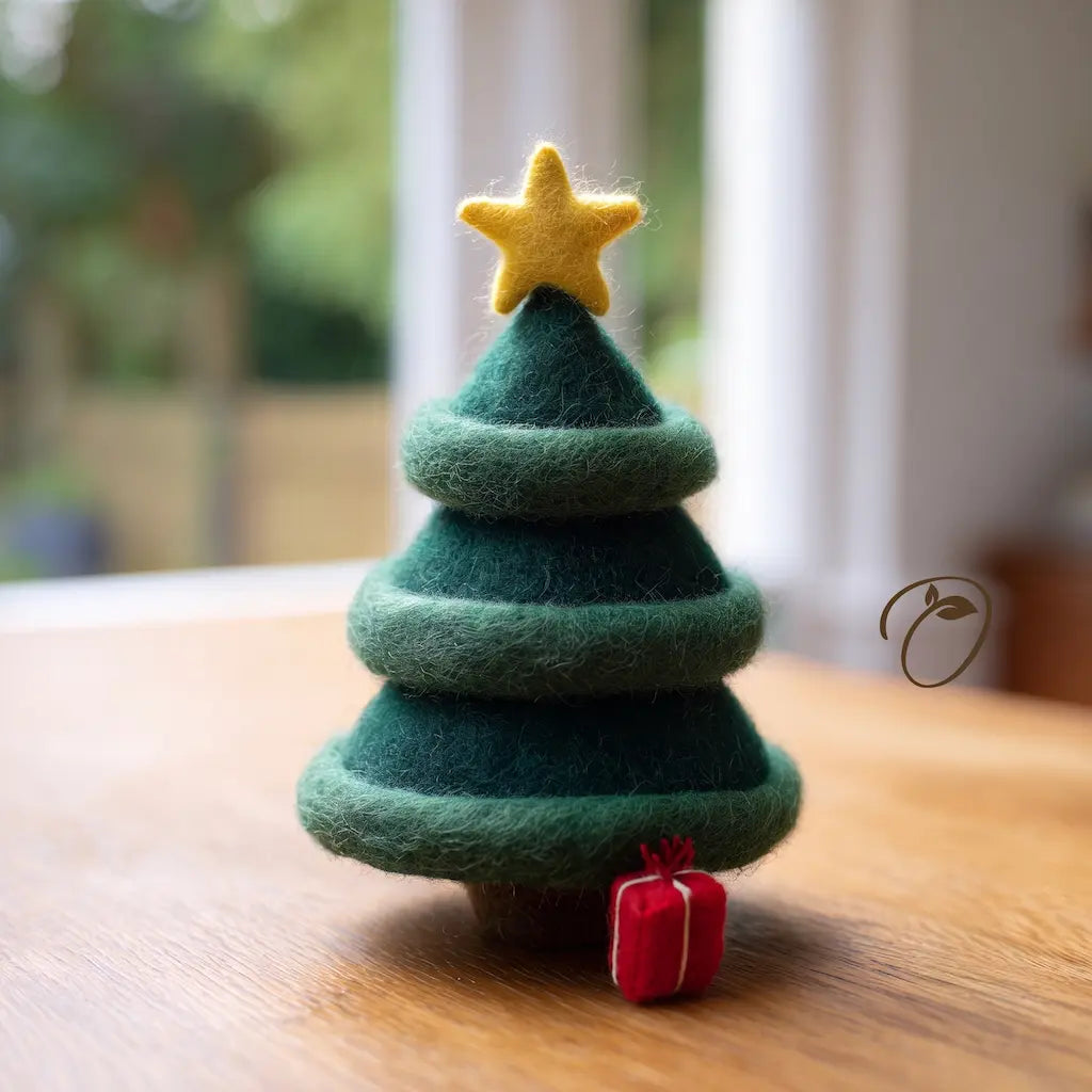 A small felt Christmas tree decoration in dark and light green with a yellow star on top and a tiny red gift box, photographed on a wooden surface with a softly blurred indoor background.
