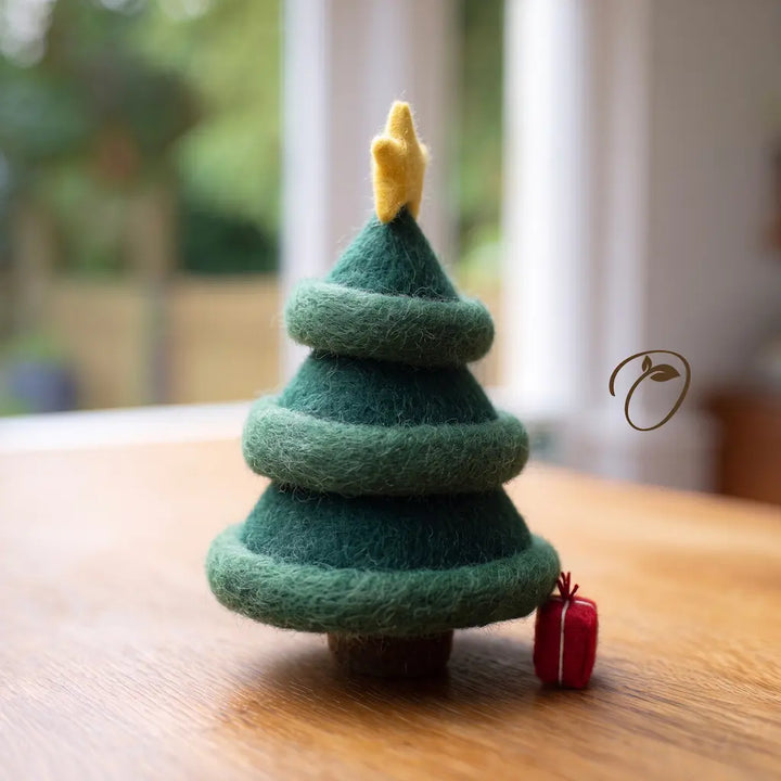 A small felt Christmas tree decoration in dark and light green with a yellow star topper, standing on a wooden table with a blurred indoor background and a tiny red gift beside it.