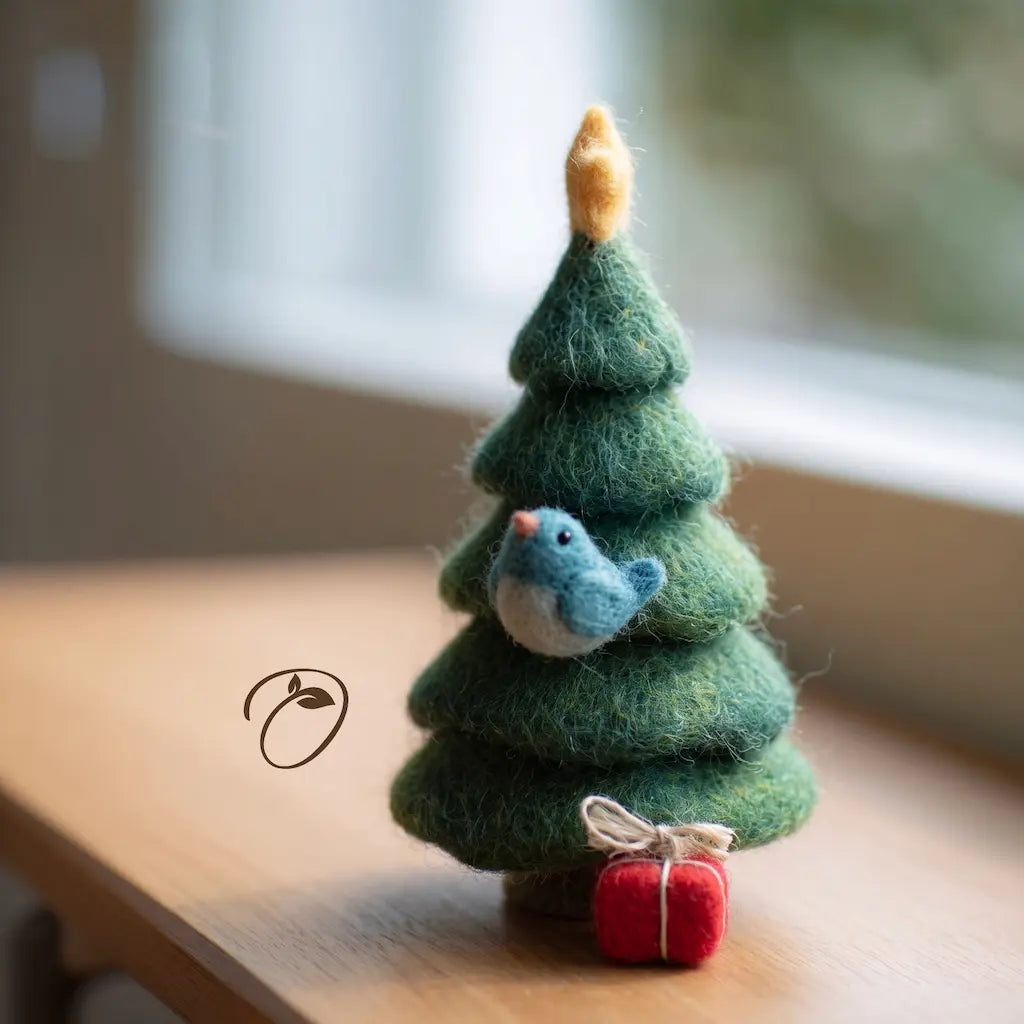 A small felt Christmas tree ornament in layered green wool, with a blue bird figure attached near the middle, a gold-tipped star on top, and a tiny red gift box at the base, photographed on a wooden surface near a window.
