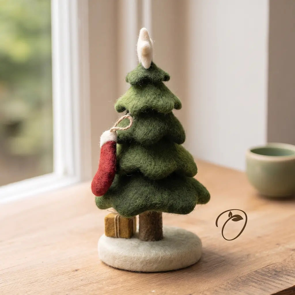 A small felt Christmas tree decoration in layered green tones with a white star topper, a red stocking hanging from the side, and a round white base, photographed on a wooden tabletop near a window.