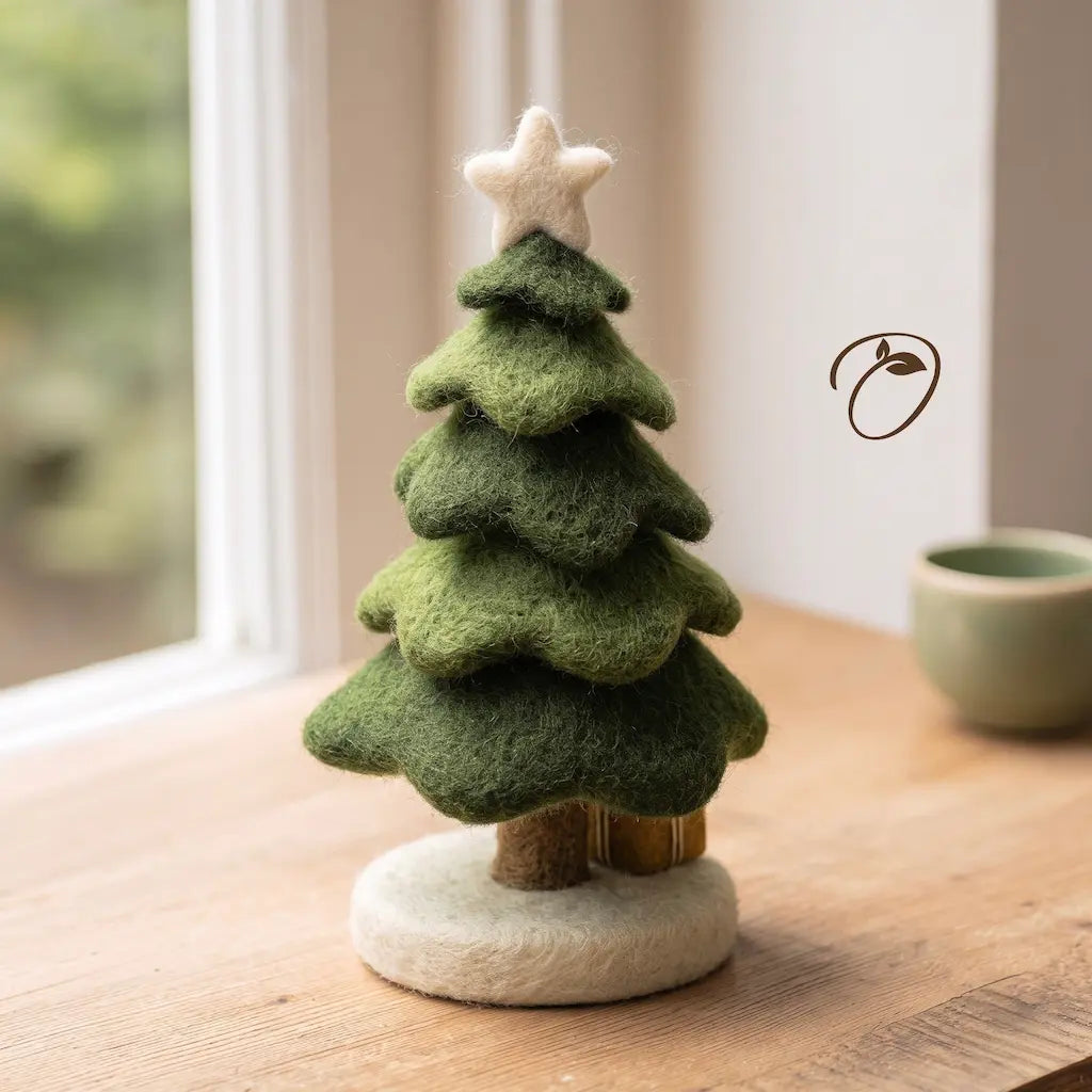 A small felt Christmas tree ornament with layered green foliage, a cream star topper, and a round cream base, displayed on a wooden tabletop near a window.