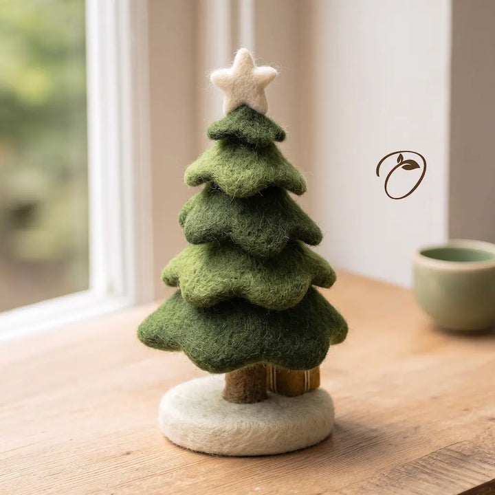 A small felt Christmas tree ornament with layered green foliage, a cream star topper, and a round cream base, displayed on a wooden tabletop near a window.