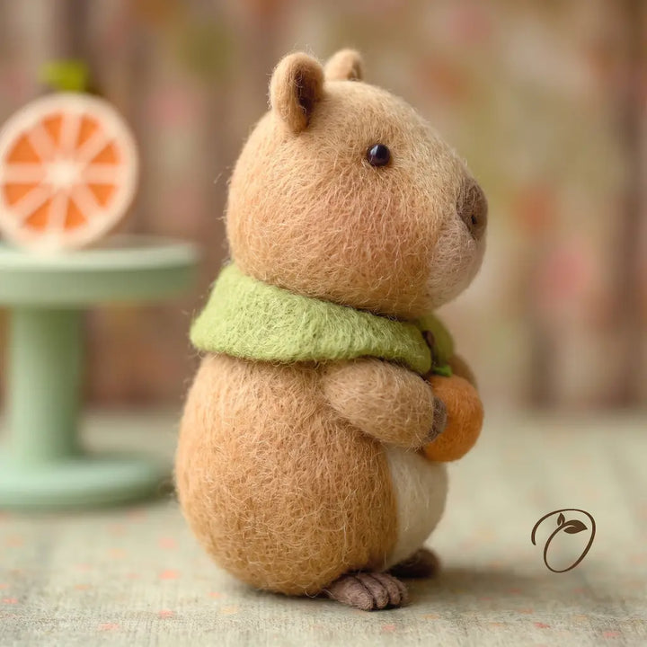 A needle-felted toy animal with brown fur, a light green scarf, and a small orange accessory, posed on a soft blurred background with a decorative stand visible at left.