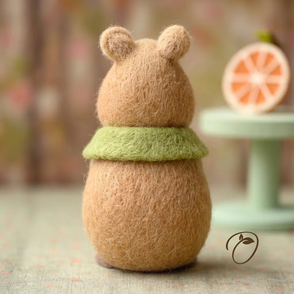 A small felted wool bear figurine with a light brown body and ears, wearing a light green collar, standing on a textured surface with a softly blurred background.