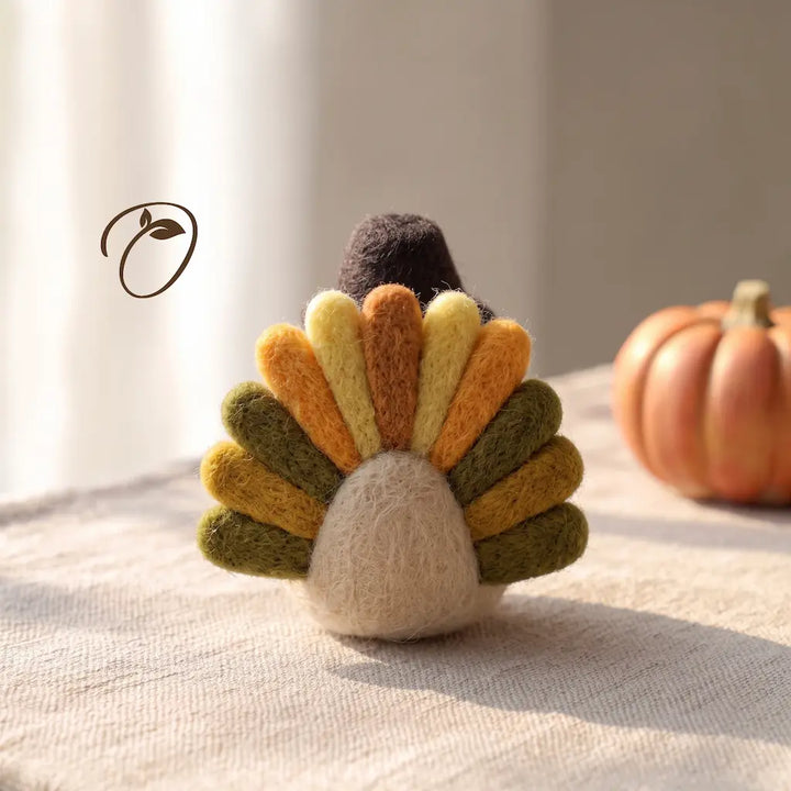 A handcrafted felt Thanksgiving turkey ornament displayed on a textured beige fabric surface with a small orange pumpkin in the background.
