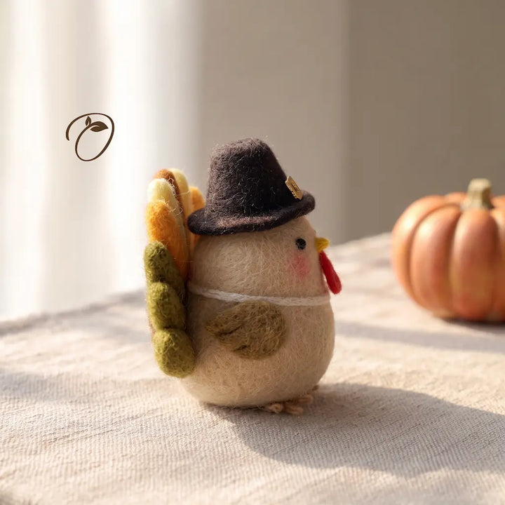 A small felt turkey decoration wearing a dark brown pilgrim hat, standing on a textured cream fabric surface with a blurred orange pumpkin in the background.