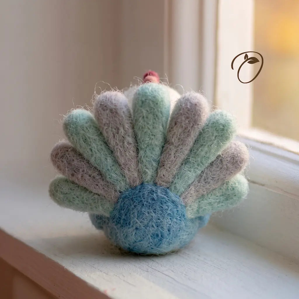 A handmade felted wool ornament shaped like a seashell, featuring pastel mint green, lavender, and light blue rays with a soft blue center and a tiny red accent on top, resting on a white windowsill.