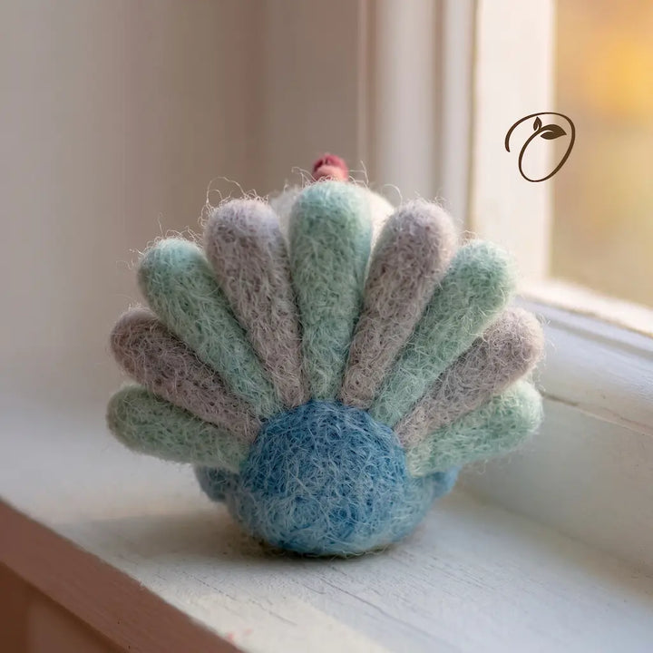 A handmade felted wool ornament shaped like a seashell, featuring pastel mint green, lavender, and light blue rays with a soft blue center and a tiny red accent on top, resting on a white windowsill.