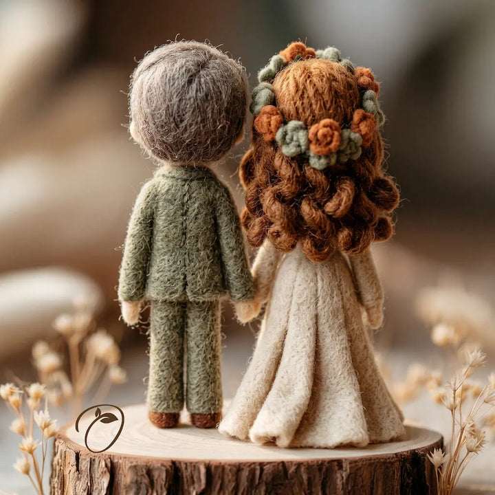 Handmade wool felt figurines of a couple seen from behind, standing on a rustic wooden log. The groom figure has short gray hair and wears a green suit, while the bride figure has long curly brown hair with a floral crown and wears a cream dress.