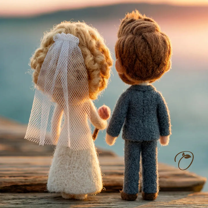 Handmade felt wedding couple figurines seen from behind, standing on a wooden dock at sunset. The bride has blonde curly hair, a white veil, and a white dress, holding hands with the groom who has brown hair and wears a blue suit.