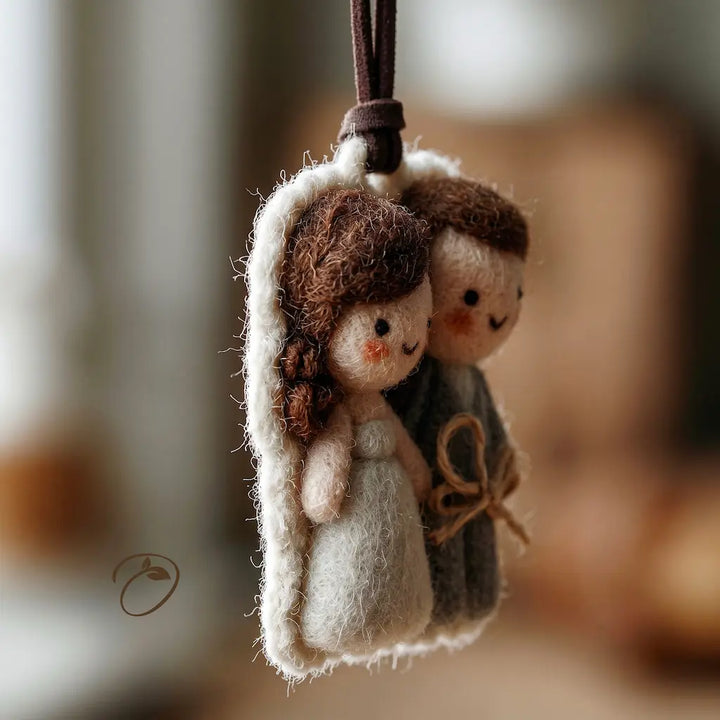 A handcrafted wool felt ornament depicting a bride and groom couple, hanging from a dark brown ribbon. The bride has brown hair and wears a white dress, while the groom has brown hair and wears a gray robe.